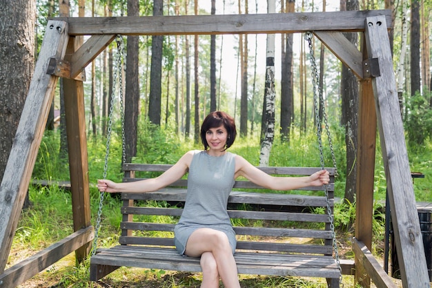 A happy brunette in a striped dress rides on a swing in a birch grove