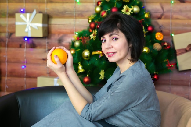 A happy brunette with a smile is sitting on the sofa in a gray dress with a bright orange in her hands against the background of a beautiful Christmas tree