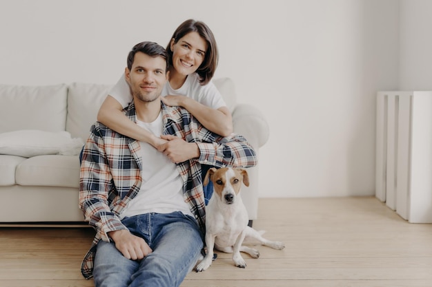 Happy brunette woman embraces husband with love being in good mood smiles positively Husband wife and dog pose together in living room of new dwelling enjoy comfort Couple in love indoor