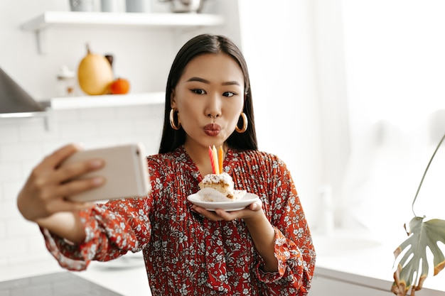 Happy brunette woman in floral red dress takes selfie and blows out candles at tasty sweet birthday cake