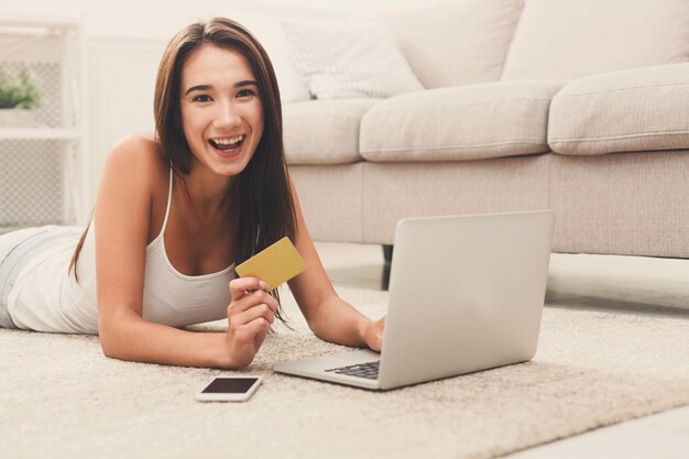 Happy brunette woman lying on the floor carpet and shopping online with credit card and laptop at home. Online shopping, technology and e-money concept