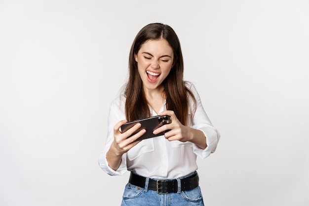 Happy brunette woman playing mobile video game smiling and looking at screen excited standing over w...