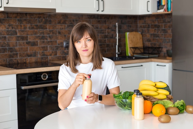 Happy brunette woman sitting with bottle of smoothie and fruits at home kitchen. Vegan meal and detox concept. Girl with white t-shirt opening fresh cocktail. Mock up