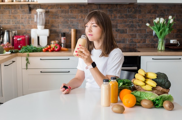 Happy brunette woman sitting with bottle of smoothie and fruits at home kitchen Vegan meal and detox concept Girl with white tshirt drinking fresh cocktail Mockup of packaging