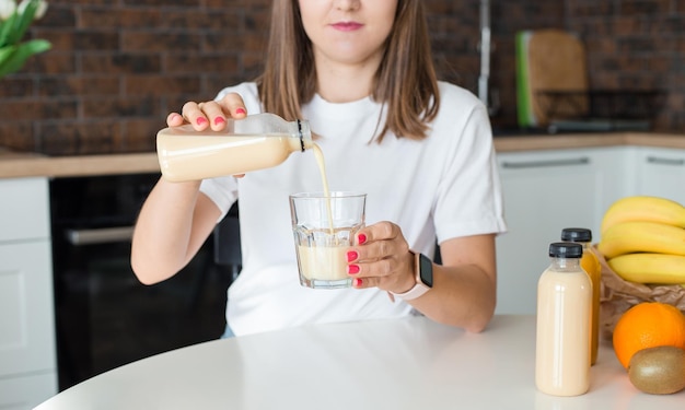 Happy brunette woman sitting with bottle of smoothie and fruits at home kitchen Vegan meal and detox concept Girl with white tshirt drinking fresh cocktail Mockup of packaging