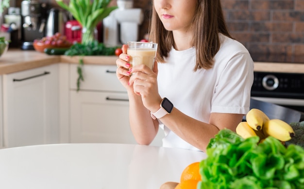 Happy brunette woman sitting with homemade glass smoothie and healthy fruits at home kitchen