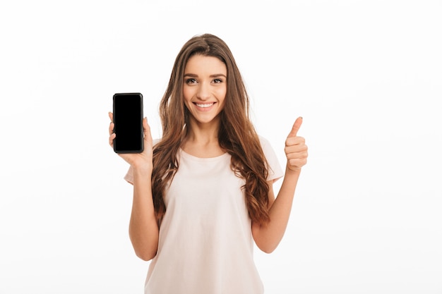 Happy brunette woman in t-shirt showing blank smartphone screen and thumb up while over white wall