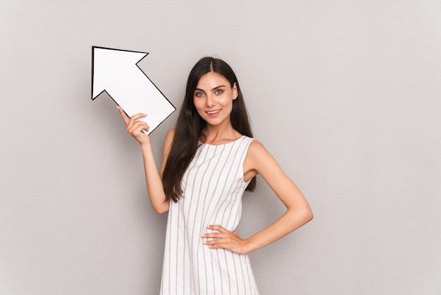  happy brunette woman wearing dress smiling and holding blank thought arrow pointer isolated over gray wall