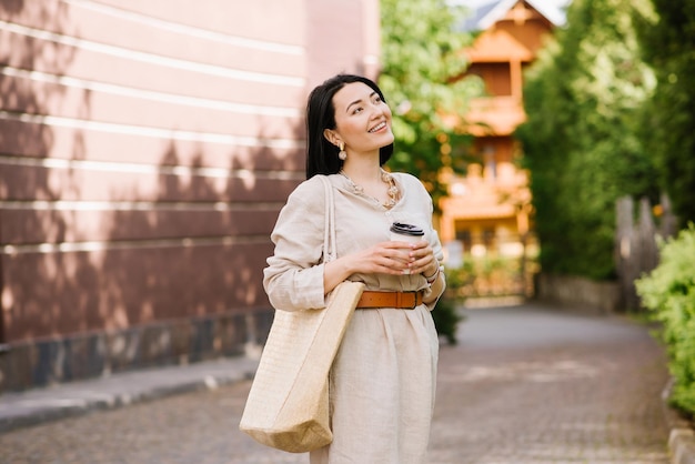 Happy brunette young woman with sunglasses and bag holding coffee walking in the city. Lifestyle portrait of smiling woman