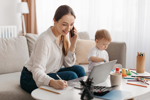 Happy business mother working at home while her little son playing next to her lady talking on phone