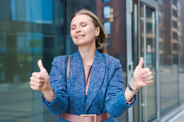 Happy business woman showing thumbs up while standing outdoors against office