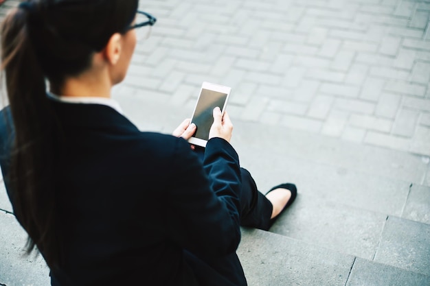 Happy business woman Smiling and cheerful woman in a business suit and glasses sitting on gray concrete steps holds the phone in her hands