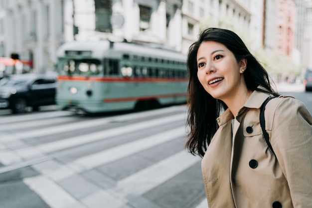 happy business woman smiling waiting for crossing zebra road with traffic bus car vehicles driving in background. young asian lady in busy business area in morning. charming college girl look around