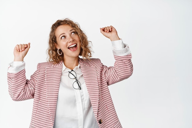 Happy business woman in suit jumping and making fist pump gesture, laughing and having fun, standing pumped up against white background