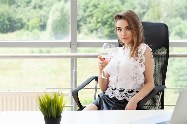 Happy business woman toasting with wine glass in office and looking at camera