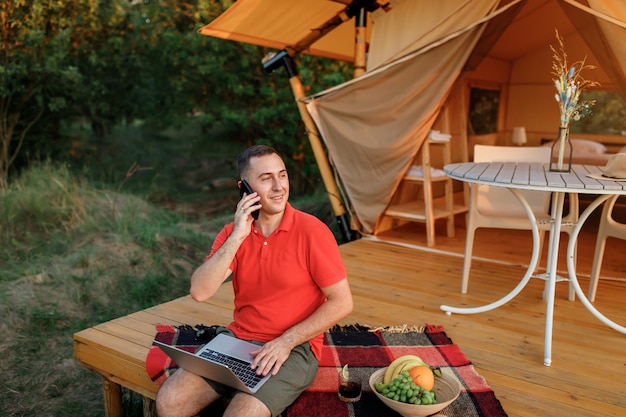 Happy businessman freelance using laptop while talking by phone on cozy glamping tent in a summer day Luxury camping tent for outdoor holiday and vacation Lifestyle concept