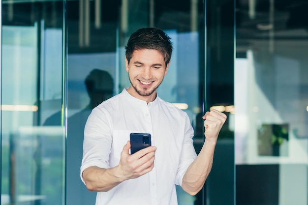 Happy businessman looking at smartphone screen making winner so gesture smiling and laughing Reading the good news celebrating achievement success