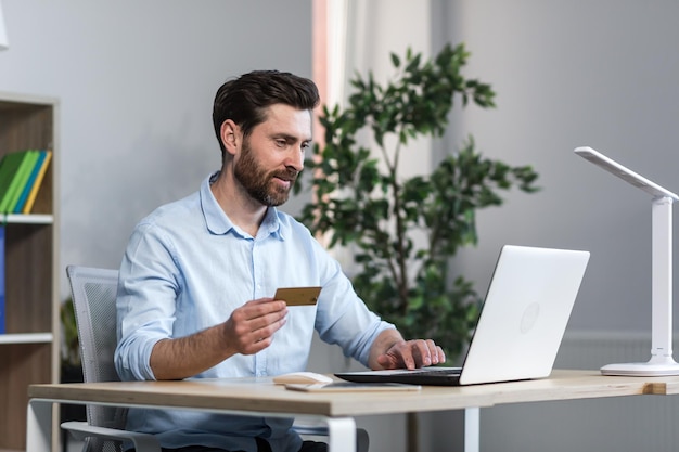 Happy businessman makes online purchases working in the office husband holds credit bank card and uses laptop