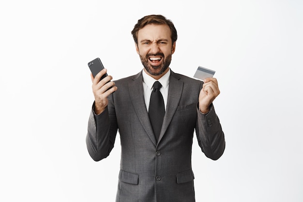 Happy businessman shouting with joy holding mobile phone and credit card winning money receive income on bank account white background