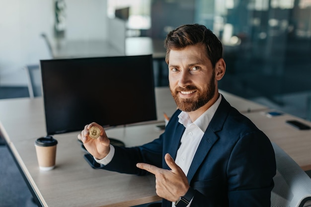 Happy businessman in suit holding golden bitcoin and pointing finger on it sitting at workplace with computer