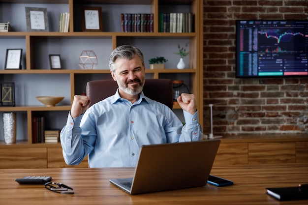 Happy businessman surprised by the good news and making yes gesture while looking at laptop sitting at office Trader enjoying success celebrating victorymake good deal get profit