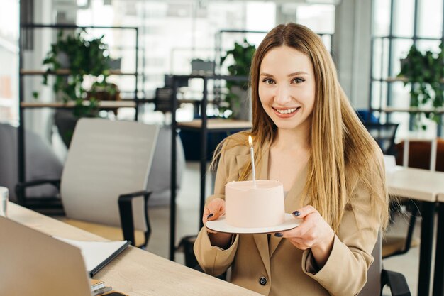 Happy businesswoman holding cake with lightened candle in the office and looking at camera