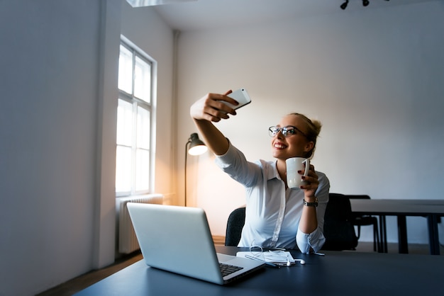 Happy businesswoman making selfie in office with cup of coffee