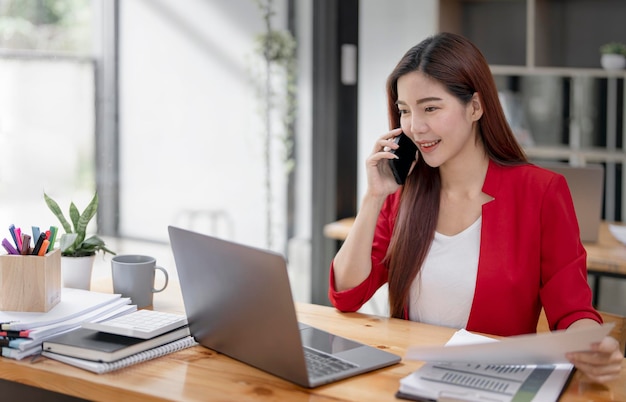 Happy businesswoman sitting at desk behind her laptop and talking with somebody on her mobile phone while working at office