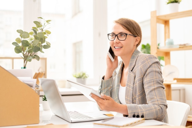 Happy businesswoman talking by smartphone and using tablet in front of laptop while sitting by desk in office