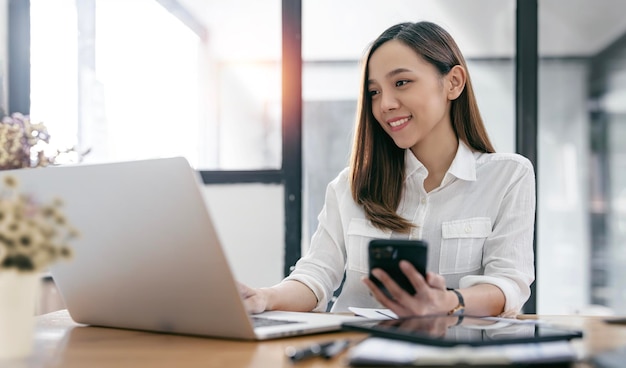 Happy businesswoman using mobile phone while analyzing weekly schedule in her notebook sitting at office desk