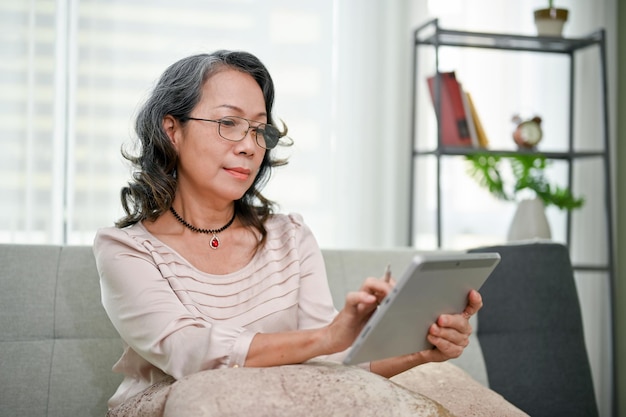 Happy and calm agedasian woman wearing glasses using tablet while chilling in living room