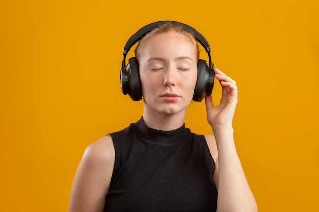 Happy and carefree excited redhead hipster girl like her new headphones, dancing with hands lifted up and smiling enjoy listening music over orange wall, boost mood with favorite song.