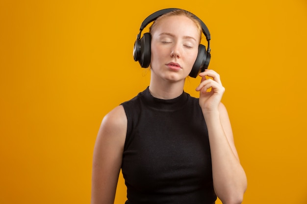 Happy and carefree excited redhead hipster girl like her new headphones, dancing with hands lifted up and smiling enjoy listening music over orange wall, boost mood with favorite song.