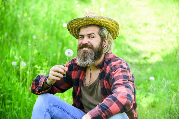 Happy and carefree life Peace of mind Rest and relax Make wish Peaceful man in straw summer hat Bearded man blowing dandelion seeds in park Mental health Peaceful guy blowing dandelion