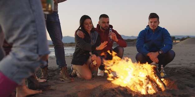 Happy Carefree Young Friends Having Fun And Drinking Beer By Bonefire On The Beach As The Sun Begins To Set