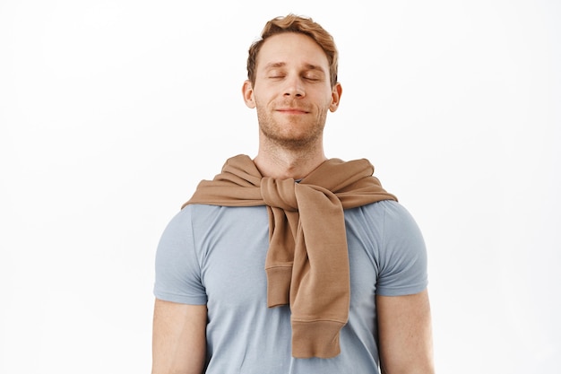 Happy and carefree young redhead man feeling healthy and pleased, close eyes and inhale fresh air, breething freely, looking relieved, standing over white wall