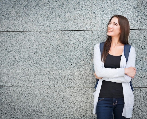 Happy casual brunette female student with backpack, staying near grey wall of an urban area. Background, copy space