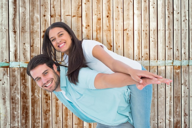 Happy casual man giving pretty girlfriend piggy back against wooden background in pale wood