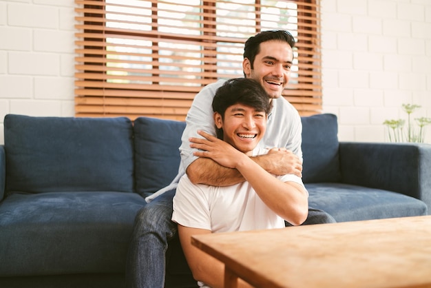 Happy caucasian and Asian LGBT couple one sitting on the sofa embracing the other sitting on the floor from the back at home Diversity of LGBT relationships A gay couple concept