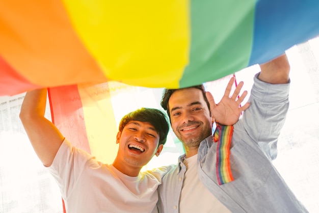 Happy caucasian and Asian LGBT couple standing and waving under the rainbow LGBT Pride flag together in the living room at home Diversity of LGBT relationships A gay couple concept