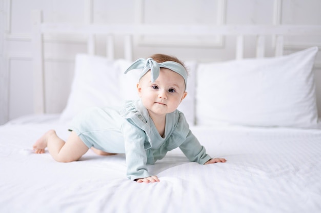 A happy Caucasian baby girl in a green bodysuit lies on the bed at home on white bedding looks at the camera laughs a healthy baby