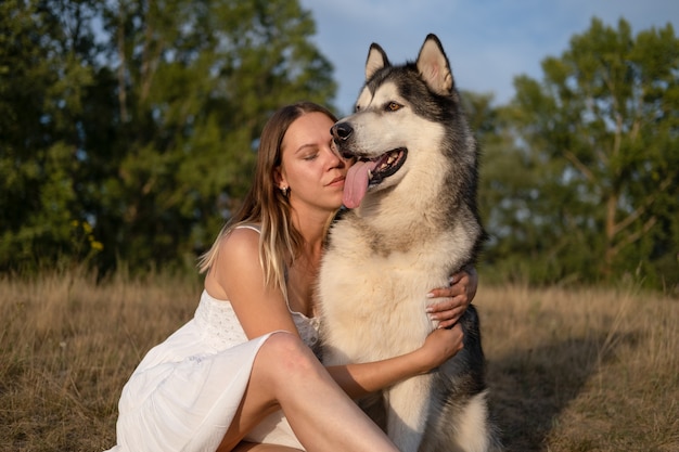 Happy Caucasian blonde woman in white dress embrace alaskan malamute dog in summer field. face to face. love and friendship between human and animal.