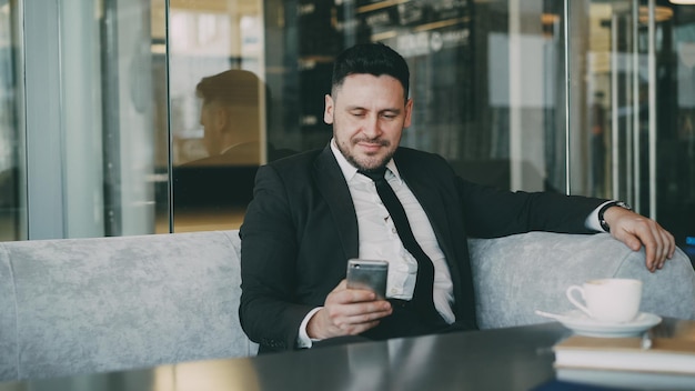 Happy Caucasian businessman in formal clothes smiling and looking at his smartphone screen while drinking coffee in airy cafe during lunch time