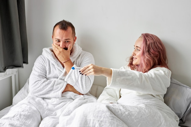 Happy caucasian couple being excited with positive pregnancy test on bed in the morning, happy reaction, man is surprised