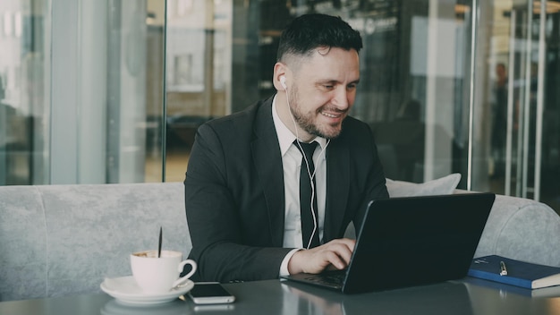 Happy Caucasian entrepreneur in formal clothes typing on his laptop while listening to music with earbuds in his ears in a modern cafe during lunch