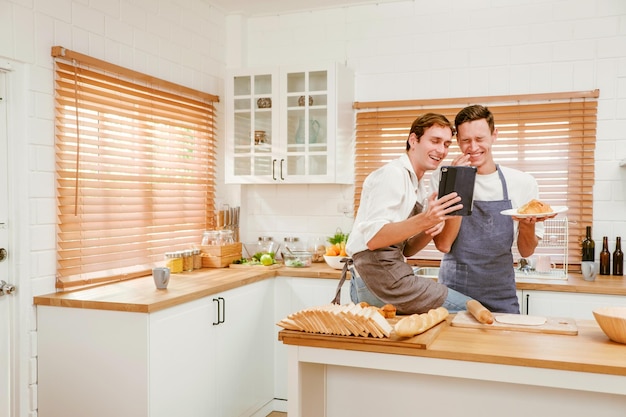 Happy Caucasian gay couple cooking together in the kitchen while watching how to cook on a digital tablet with fun and a smile Man standing by boyfriend preparing food at home LGBT couple concept