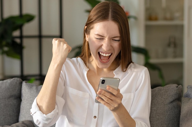 Happy caucasian millennial woman getting good news notification via mobile phone, celebrating success sitting on the couch at home