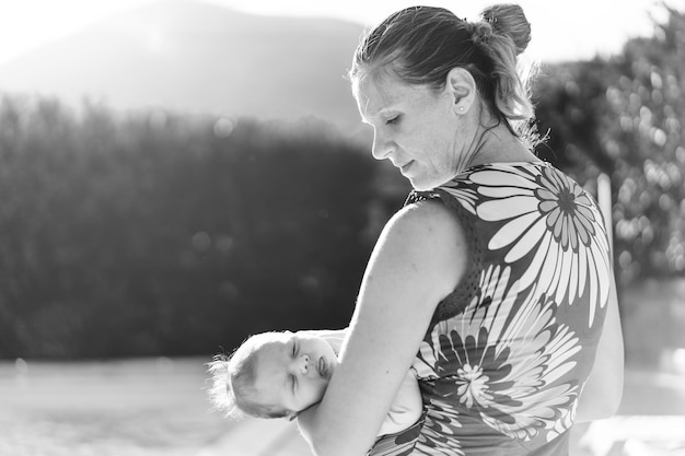 Happy caucasian mother with imperfect skin holding her baby girl in the resort garden near the swimming pool Family happiness real people and love concept Black and white