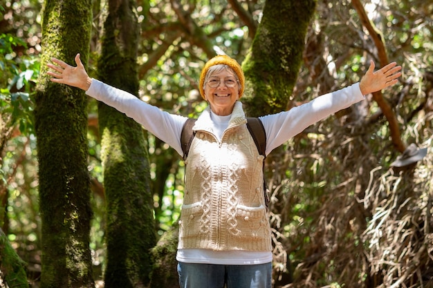 Happy caucasian senior woman hiking in the forest holding backpack enjoying freedom and nature elderly lady with eyeglasses and outstretched arms