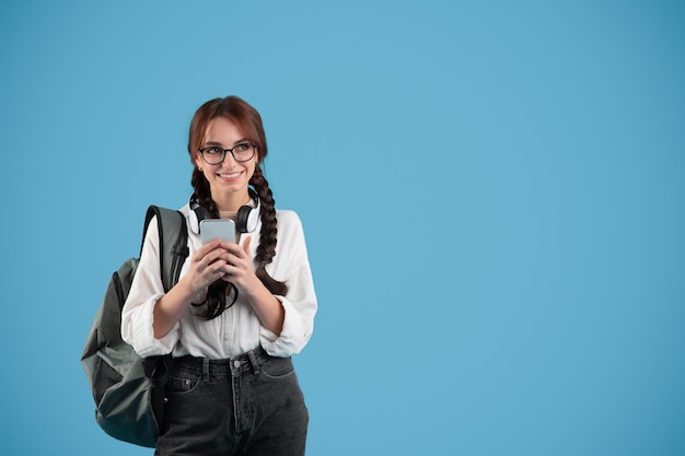 Happy caucasian teenager girl with pigtails in glasses with headphones student typing on smartphone